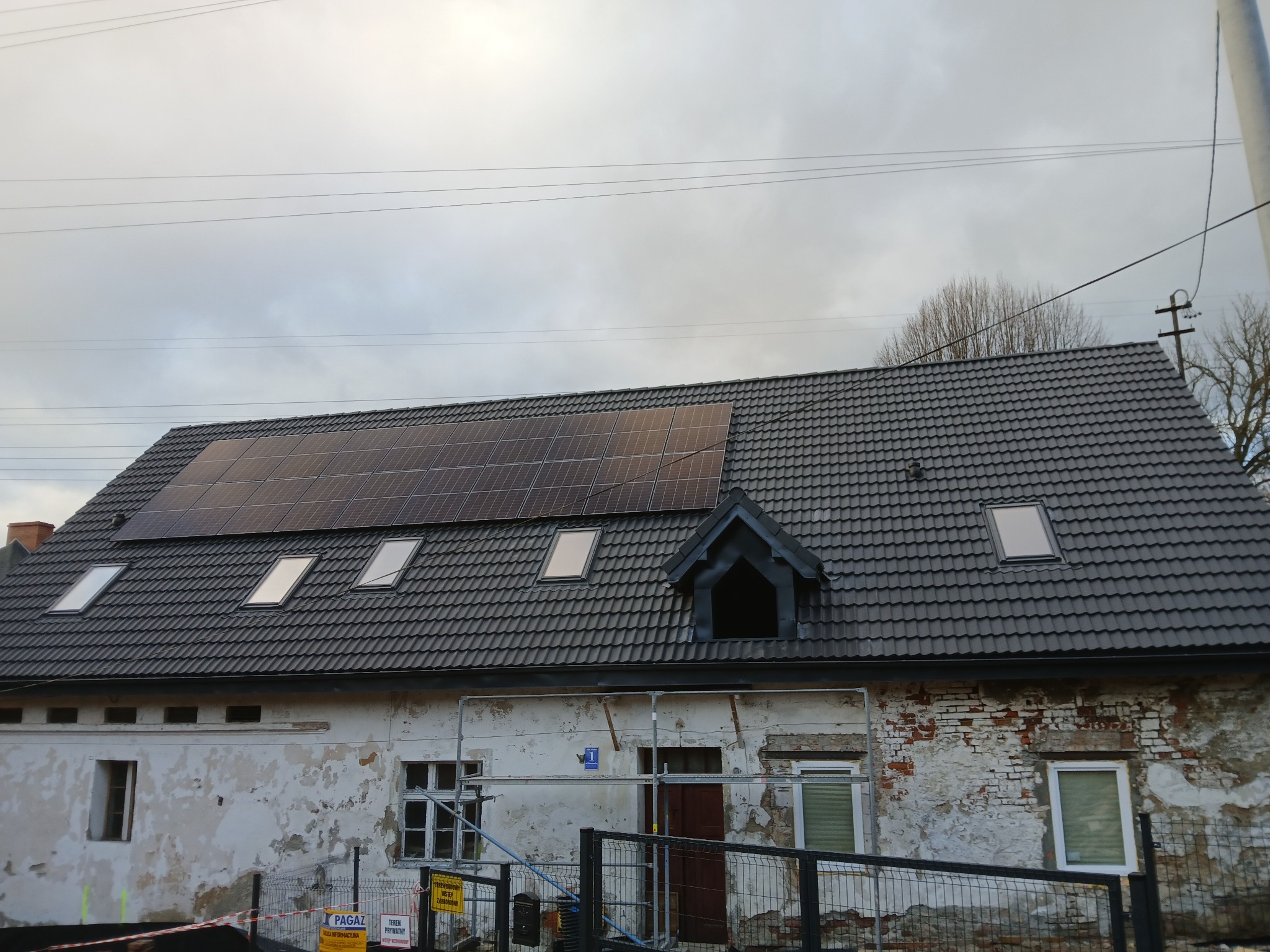Dom z szarą dachówką, panelami słonecznymi i oknami dachowymi, elewacja częściowo odnowiona, widoczne rusztowania i tablice informacyjne.