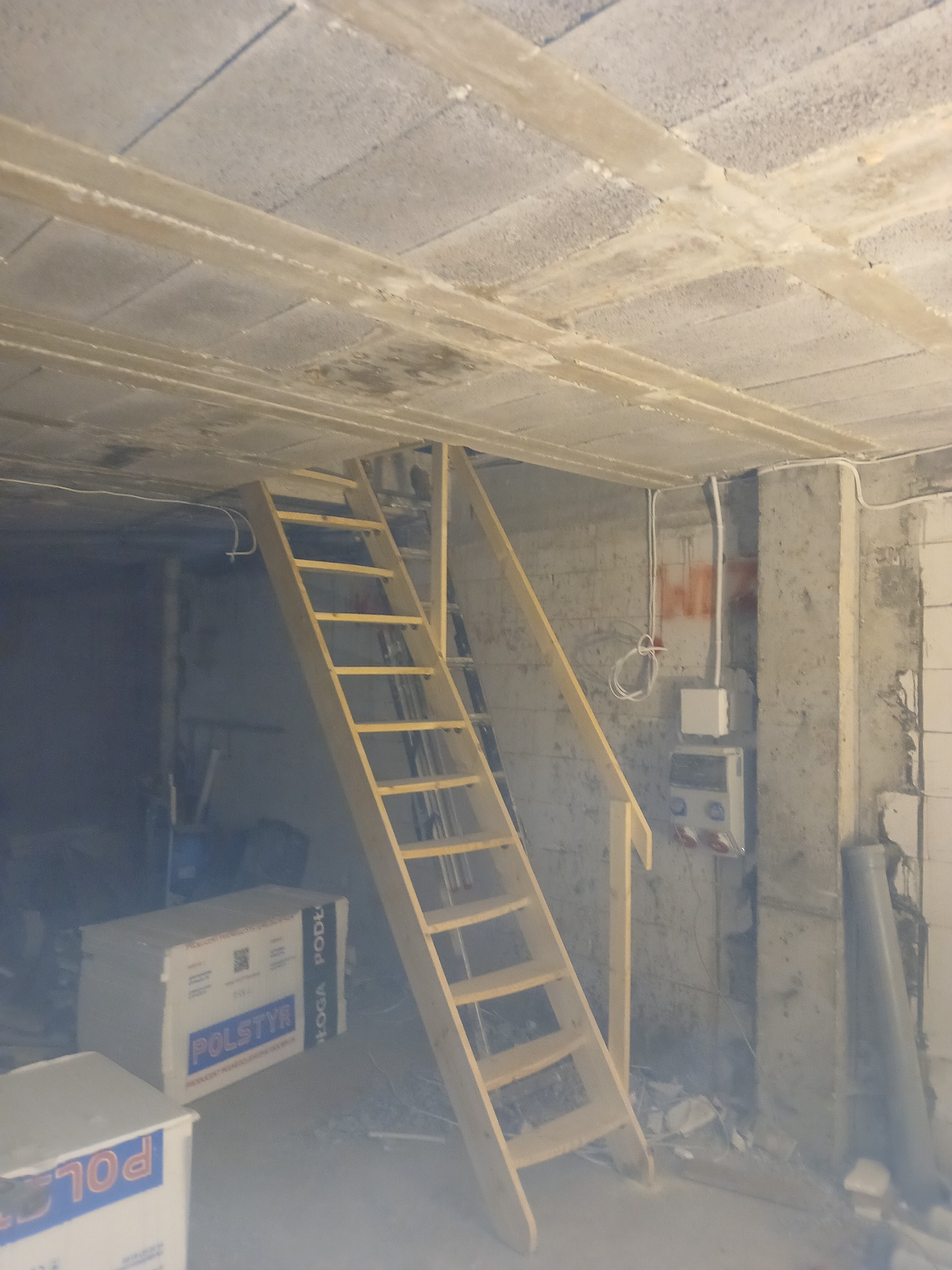 Surowe wnętrze z betonowym sufitem i ścianami, drewniane schody strychowe prowadzące do góry, widoczne kable elektryczne i puszki instalacyjne, obok schodów leżą opakowania styropianu.