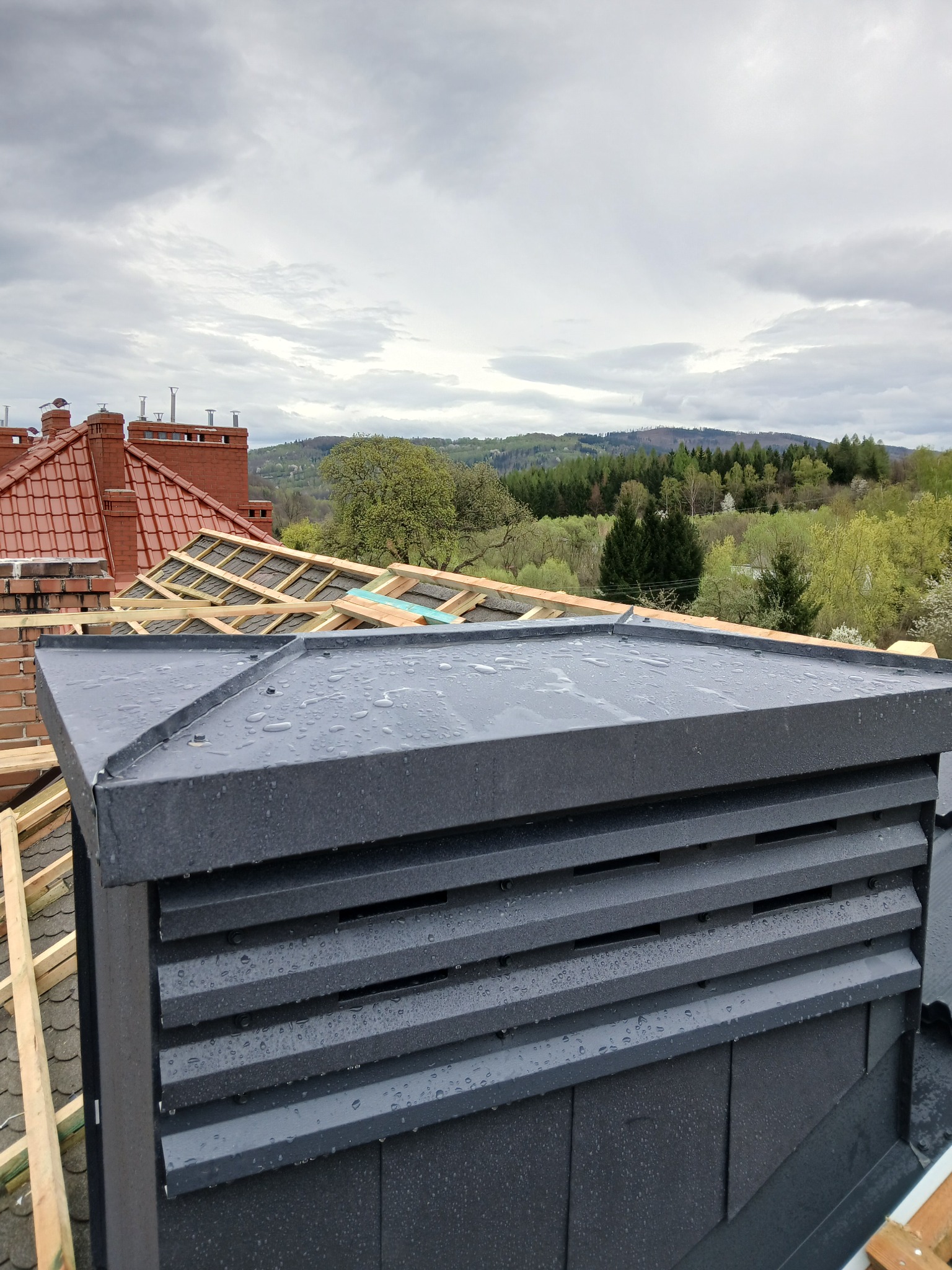 Szary, metalowy element dachu z widocznymi poziomymi żłobieniami, na tle drewnianej konstrukcji dachu i oddalonych wzgórz, w pochmurny dzień po deszczu.