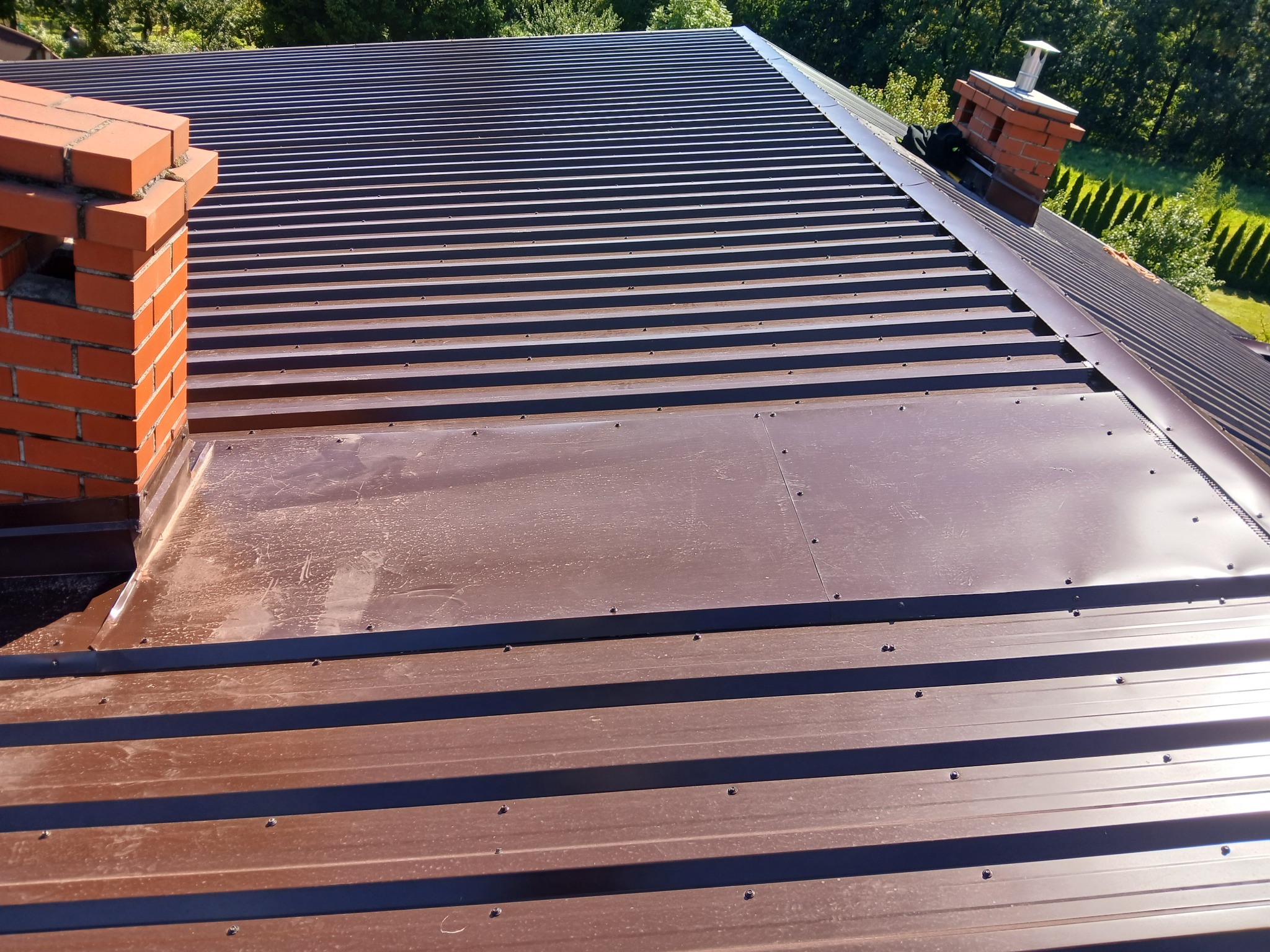 Nowe, brązowe pokrycie dachu z blachy trapezowej, widoczne kominy z cegły. Dach połaciowy z wyraźnymi liniami, świeżo po montażu, na tle zieleni drzew.