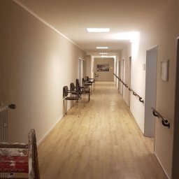 Col-Han - Długi korytarz w budynku użyteczności publicznej, świeżo po odnowieniu, z jasnymi ścianami, drewnopodobną podłogą i schodami malarskimi w lewym dolnym rogu.