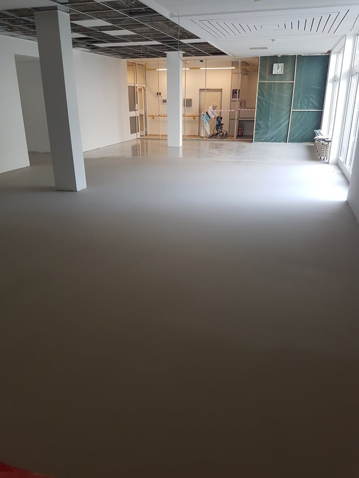 Wnętrze biura w trakcie prac wykończeniowych: świeżo wylana, gładka posadzka samopoziomująca, widoczne filary, częściowo zabudowane ściany, pracownik w kombinezonie ochronnym z wózkiem w oddali.