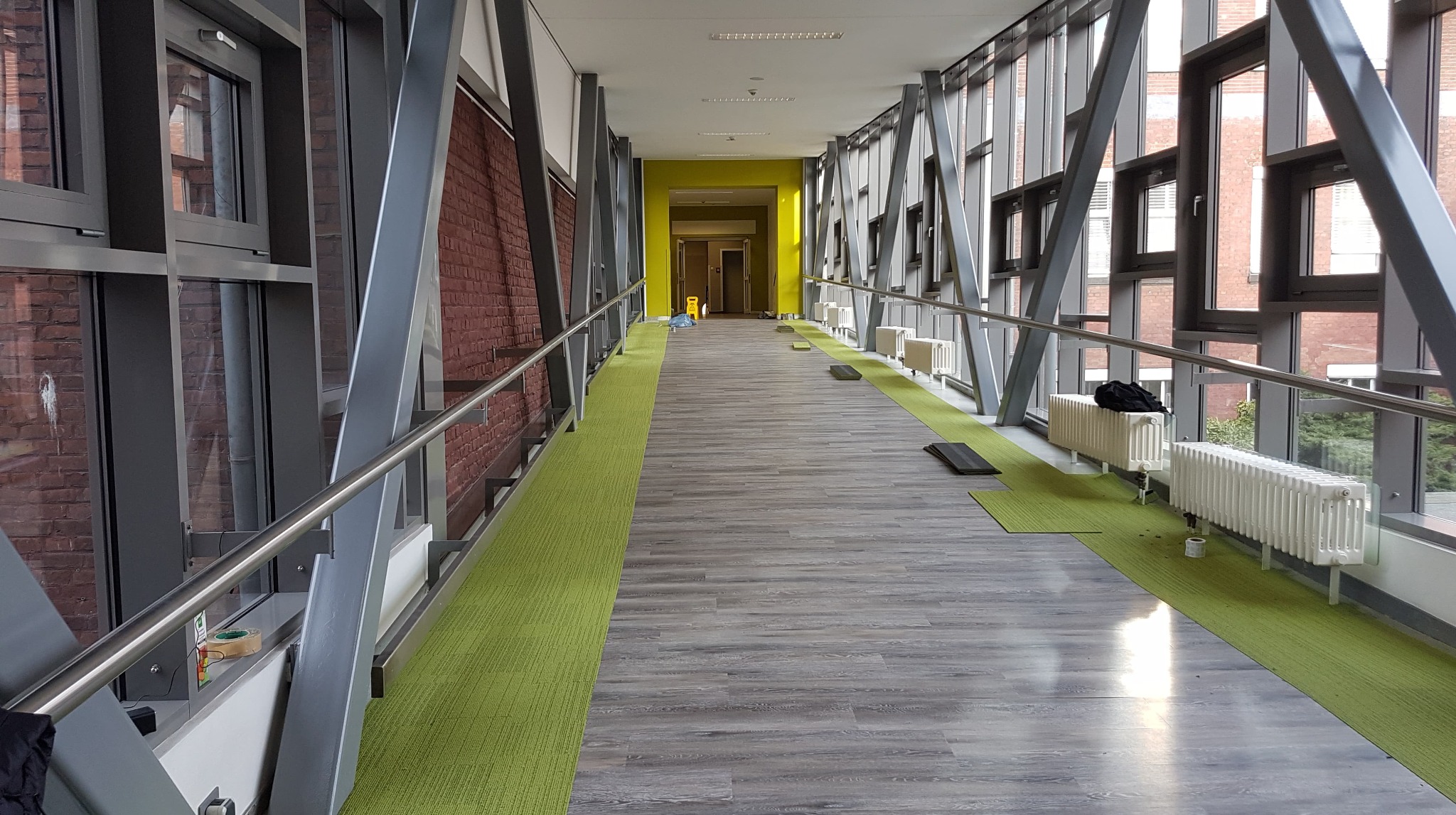 Korytarz w budynku biurowym z szarymi panelami podłogowymi imitującymi drewno i zielonymi wykładzinami dywanowymi wzdłuż ścian, widoczne grzejniki i przygotowane panele do położenia.