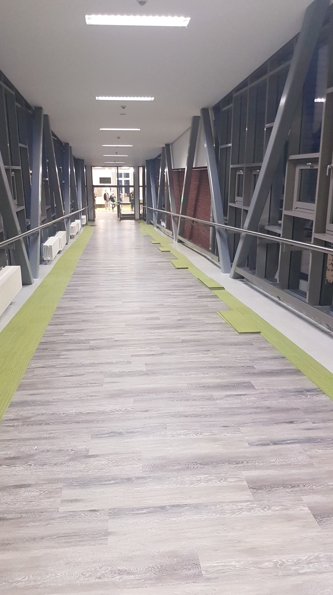 Korytarz z szarymi panelami podłogowymi imitującymi drewno i zielonymi wykładzinami dywanowymi po bokach, w tle widoczne metalowe konstrukcje i grzejniki.