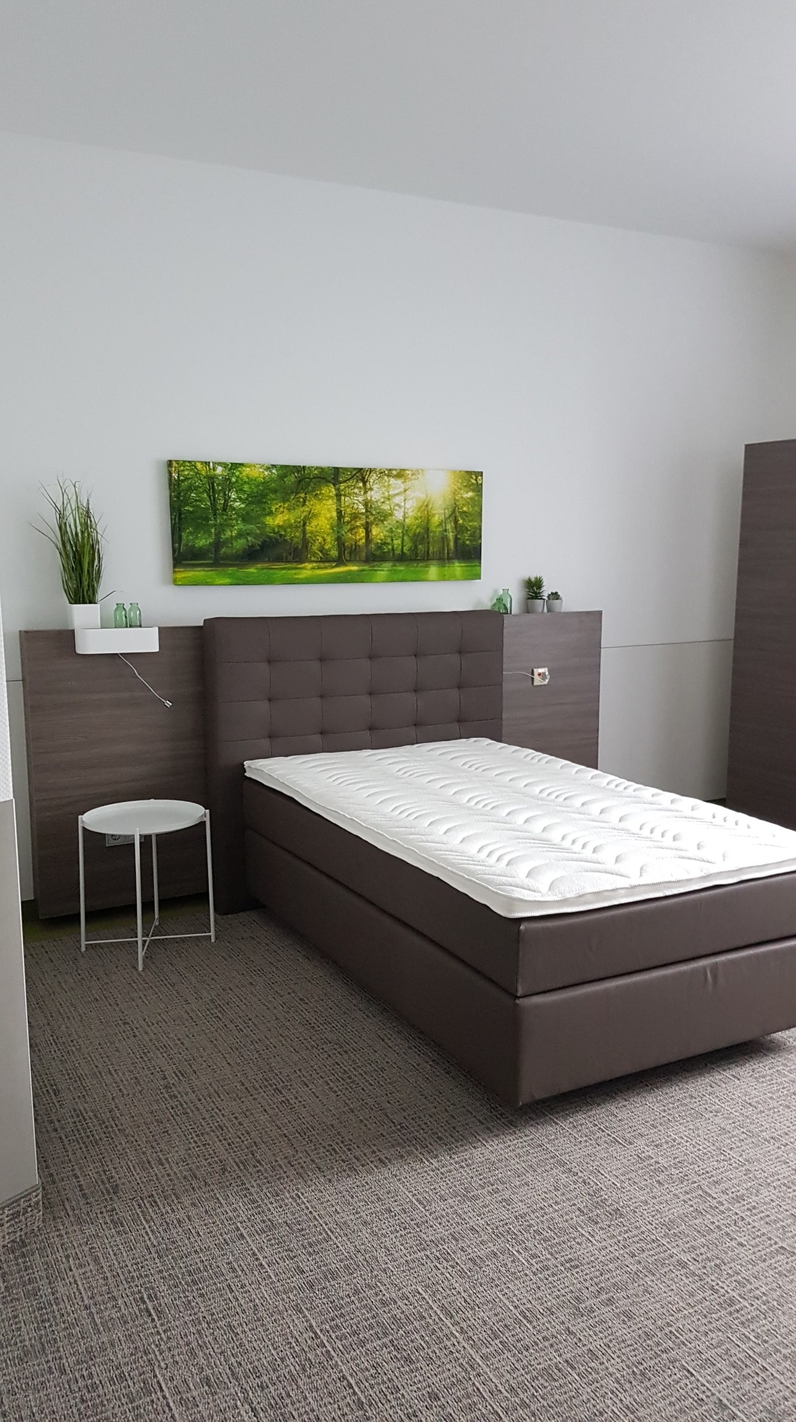 Pokój z brązowym łóżkiem kontynentalnym, tapicerowanym zagłówkiem, stolikiem nocnym i obrazem leśnym na ścianie.