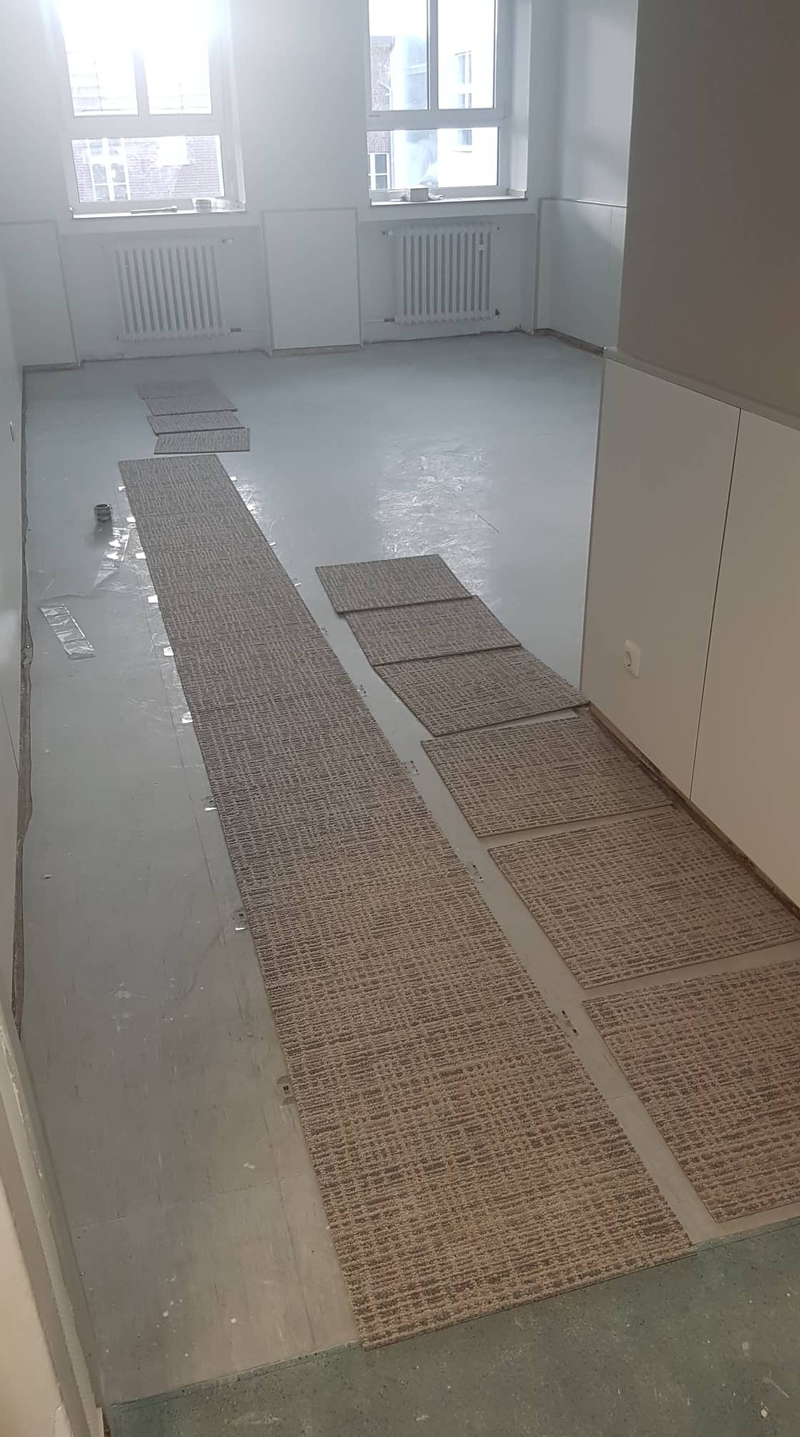 Przygotowanie do położenia wykładziny dywanowej w jasnym pomieszczeniu biurowym, widoczne paski i kwadraty wykładziny ułożone na podłodze, zabezpieczone taśmą. Radiatory pod oknami.