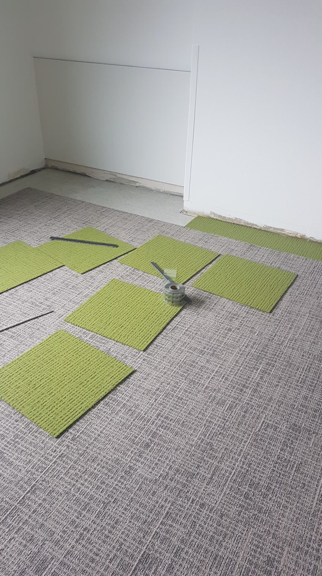 Układanie zielonych kwadratowych płytek dywanowych na szarym dywanie w pomieszczeniu z białymi ścianami, widoczne narzędzia montażowe.