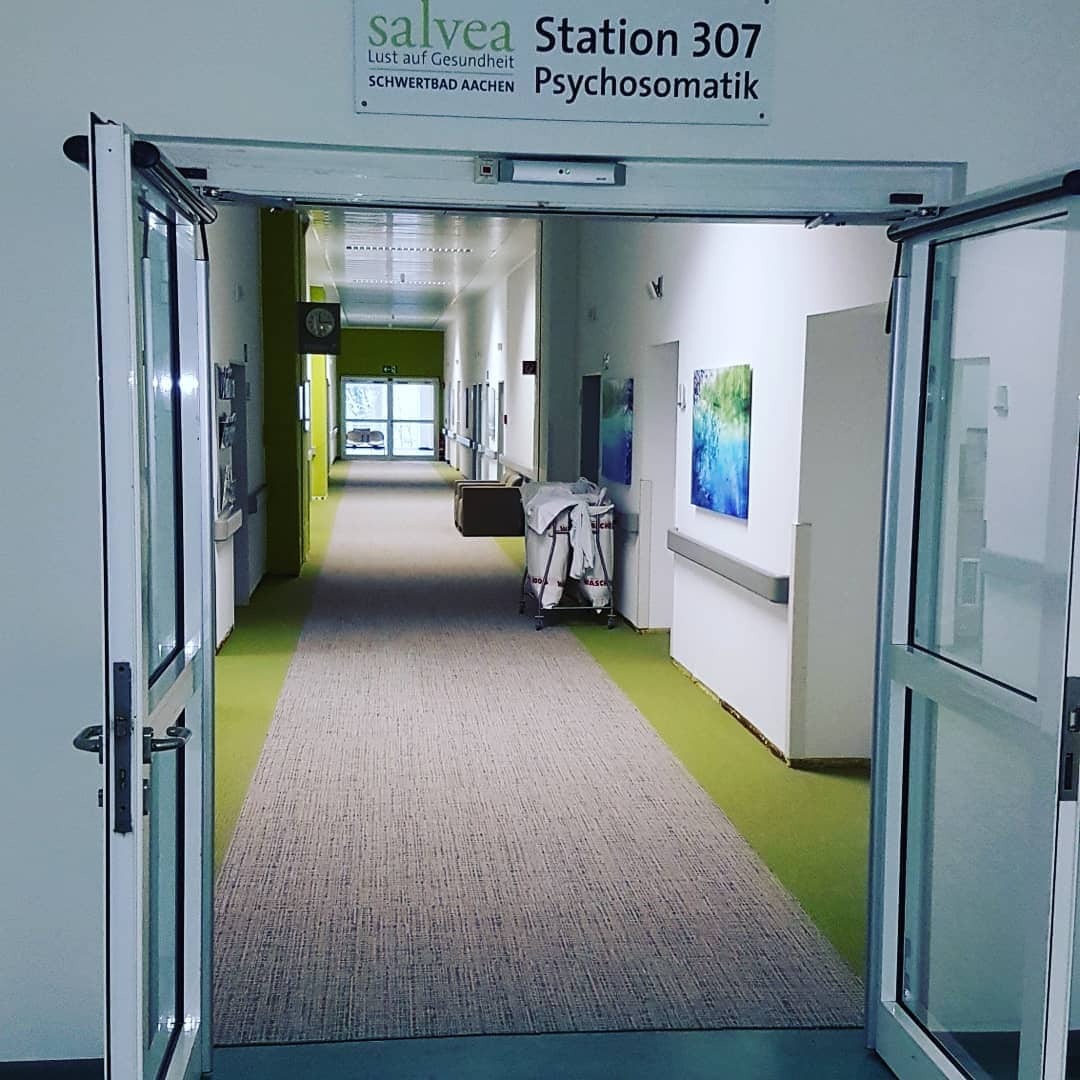 Długi korytarz w placówce medycznej, z otwartymi szklanymi drzwiami po obu stronach, jasne ściany, zielony pas na podłodze po bokach dywanu, w oddali wózek z pościelą oraz drzwi na końcu korytarza.