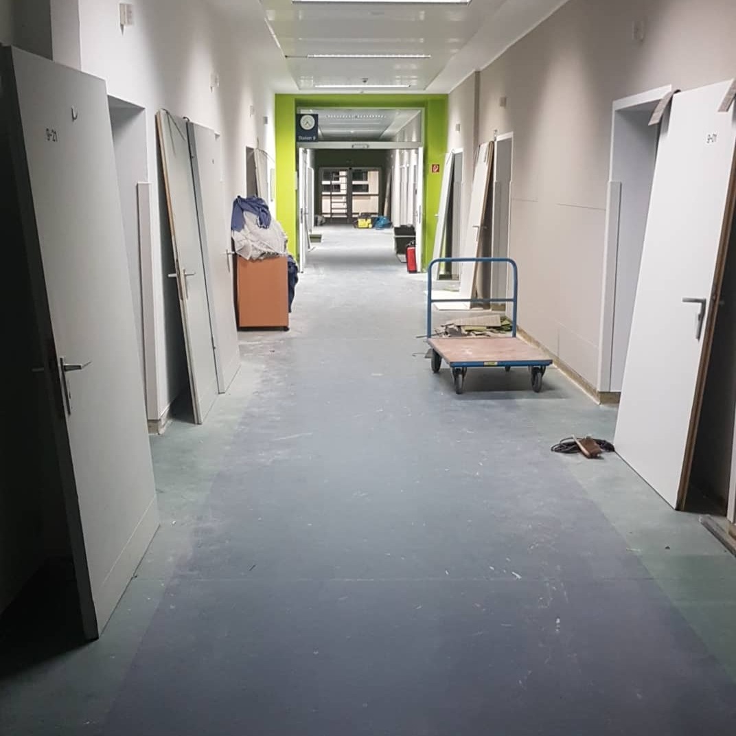 Korytarz w trakcie prac wykończeniowych: ściany przygotowane pod malowanie, nowe białe drzwi opierają się o ściany, w tle widoczny wózek transportowy i pomalowana na zielono rama drzwi.