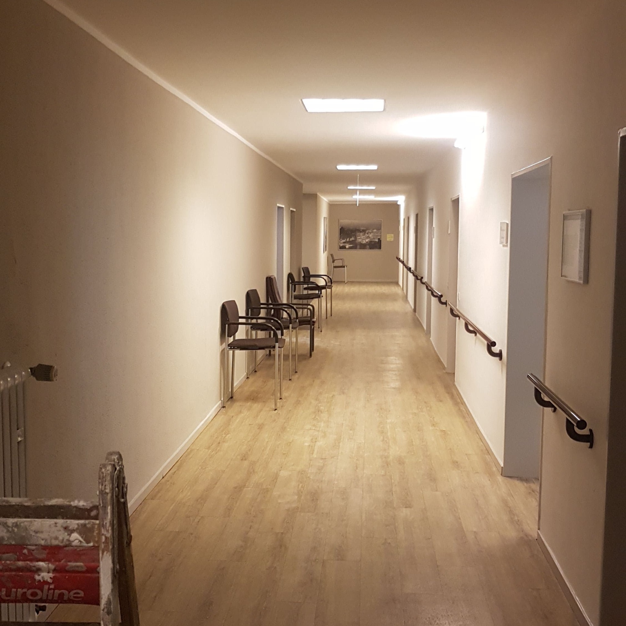Długi korytarz w budynku użyteczności publicznej, świeżo po odnowieniu, z jasnymi ścianami, drewnopodobną podłogą i schodami malarskimi w lewym dolnym rogu.