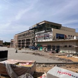 Marwikt Usługi Budowlane - Budynek komercyjny w trakcie prac elewacyjnych, widoczne rusztowania, podnośnik koszowy z pracownikami, palety z materiałem izolacyjnym Rockwool oraz częściowo otynkowana fasada.