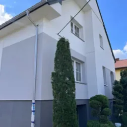 Dwukolorowa elewacja domu jednorodzinnego w odcieniach szarości i bieli, z widoczną rynną zabezpieczoną folią ochronną oraz iglakiem w pierwszym planie.