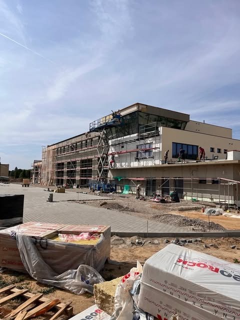 Budynek komercyjny w trakcie prac elewacyjnych, widoczne rusztowania, podnośnik koszowy z pracownikami, palety z materiałem izolacyjnym Rockwool oraz częściowo otynkowana fasada.