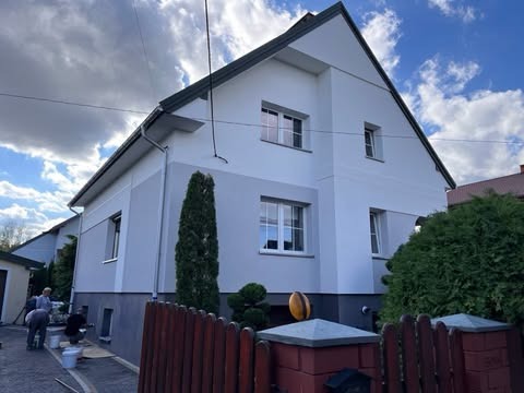 Dwukolorowa elewacja domu jednorodzinnego po dociepleniu, widoczne prace wykończeniowe przy cokole i pracownicy w trakcie malowania, Tomaszów Mazowiecki.