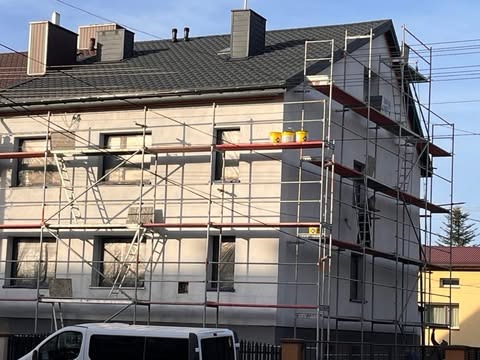 Dwupiętrowy dom w trakcie prac elewacyjnych, rusztowania rozstawione, widoczne trzy żółte wiadra na rusztowaniu, okna zabezpieczone folią.