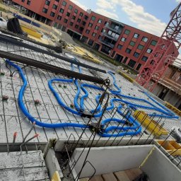 FHDC Firma Handlowa Dawid Czyż - Betonowy strop w trakcie budowy z widocznymi niebieskimi rurami instalacji wentylacyjnej, zbrojeniem i szalunkami, w tle budynek mieszkalny i żurawie budowlane.