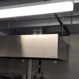 Kuchenny okap przemysłowy ze stali nierdzewnej marki Technica, zamontowany w pomieszczeniu z szarymi płytkami, widoczne kanały wentylacyjne pod sufitem i oświetlenie jarzeniowe.