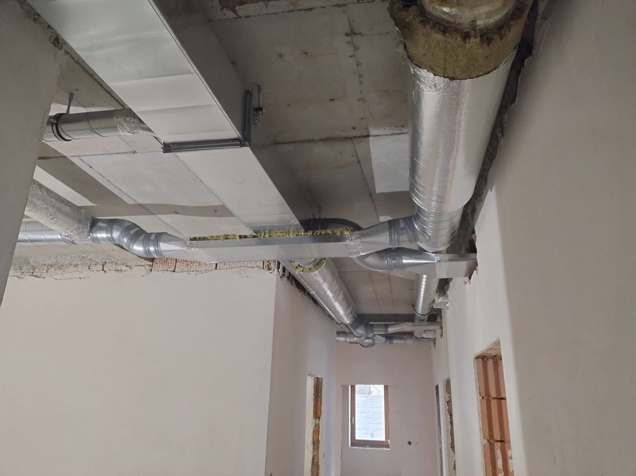 System kanałów wentylacyjnych, częściowo izolowanych, zamontowany pod surowym betonowym stropem w budynku w trakcie wykańczania, widoczne okno i fragment ściany z cegieł.