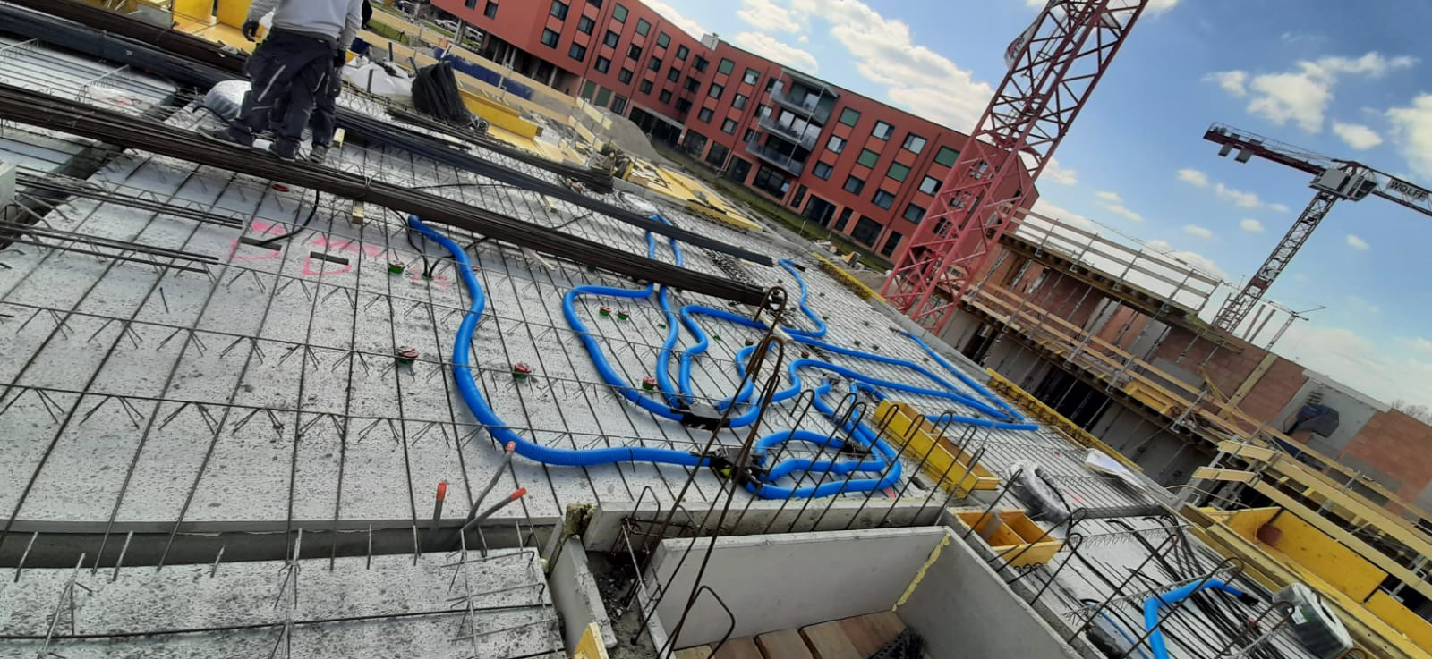 Betonowy strop w trakcie budowy z widocznymi niebieskimi rurami instalacji wentylacyjnej, zbrojeniem i szalunkami, w tle budynek mieszkalny i żurawie budowlane.