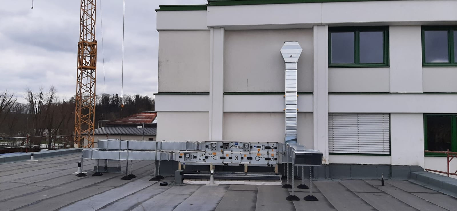 Instalacja kanałów wentylacyjnych na dachu budynku komercyjnego, widoczny pionowy kanał wentylacyjny przy ścianie, poziome kanały oraz centrala wentylacyjna na podporach, w tle żuraw budowlany...