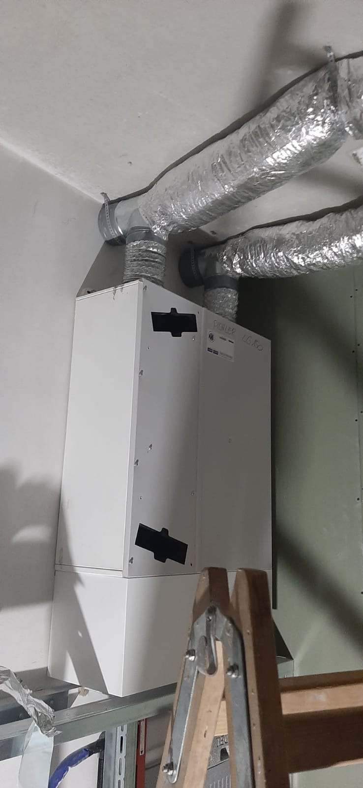 Biała jednostka centralna wentylacyjna z podłączonymi przewodami wentylacyjnymi aluminiowymi, zamontowana przy ścianie, widoczna drabina.