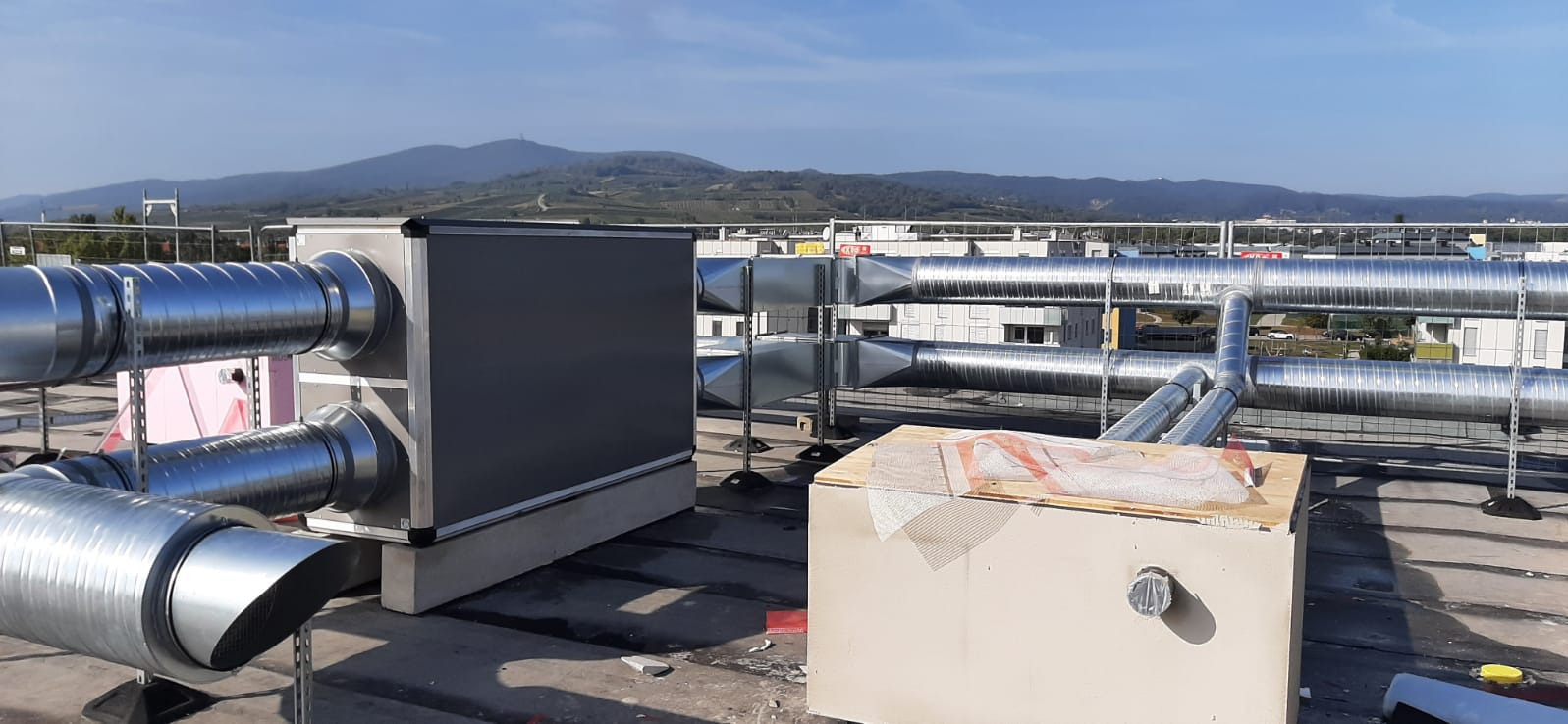 System wentylacji z metalowymi rurami na dachu budynku komercyjnego, w tle góry i budynki.