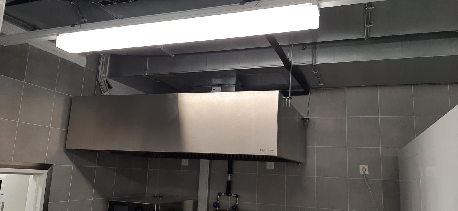 Kuchenny okap przemysłowy ze stali nierdzewnej marki Technica, zamontowany w pomieszczeniu z szarymi płytkami, widoczne kanały wentylacyjne pod sufitem i oświetlenie jarzeniowe.
