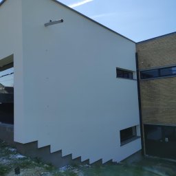 Building Industry - Nowoczesny budynek z białą elewacją i elementami z cegły, widoczne detale architektoniczne, takie jak schodkowa podstawa ściany i metalowa rura wentylacyjna, na tle błękitnego nieba z smugą...