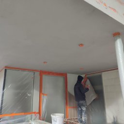 Building Industry - Pracownik w trakcie nakładania warstwy wykończeniowej na elewację domu jednorodzinnego, widoczne zabezpieczone okna folią i taśmą, worek z zaprawą Polystar oraz wiadro z farbą.