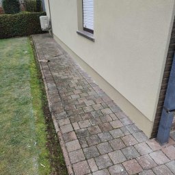 Building Industry - Fragment elewacji budynku z beżowym tynkiem, widoczne okno z roletą, klimatyzator na ścianie oraz chodnik z kostki brukowej. Na trawniku widoczne narzędzia.