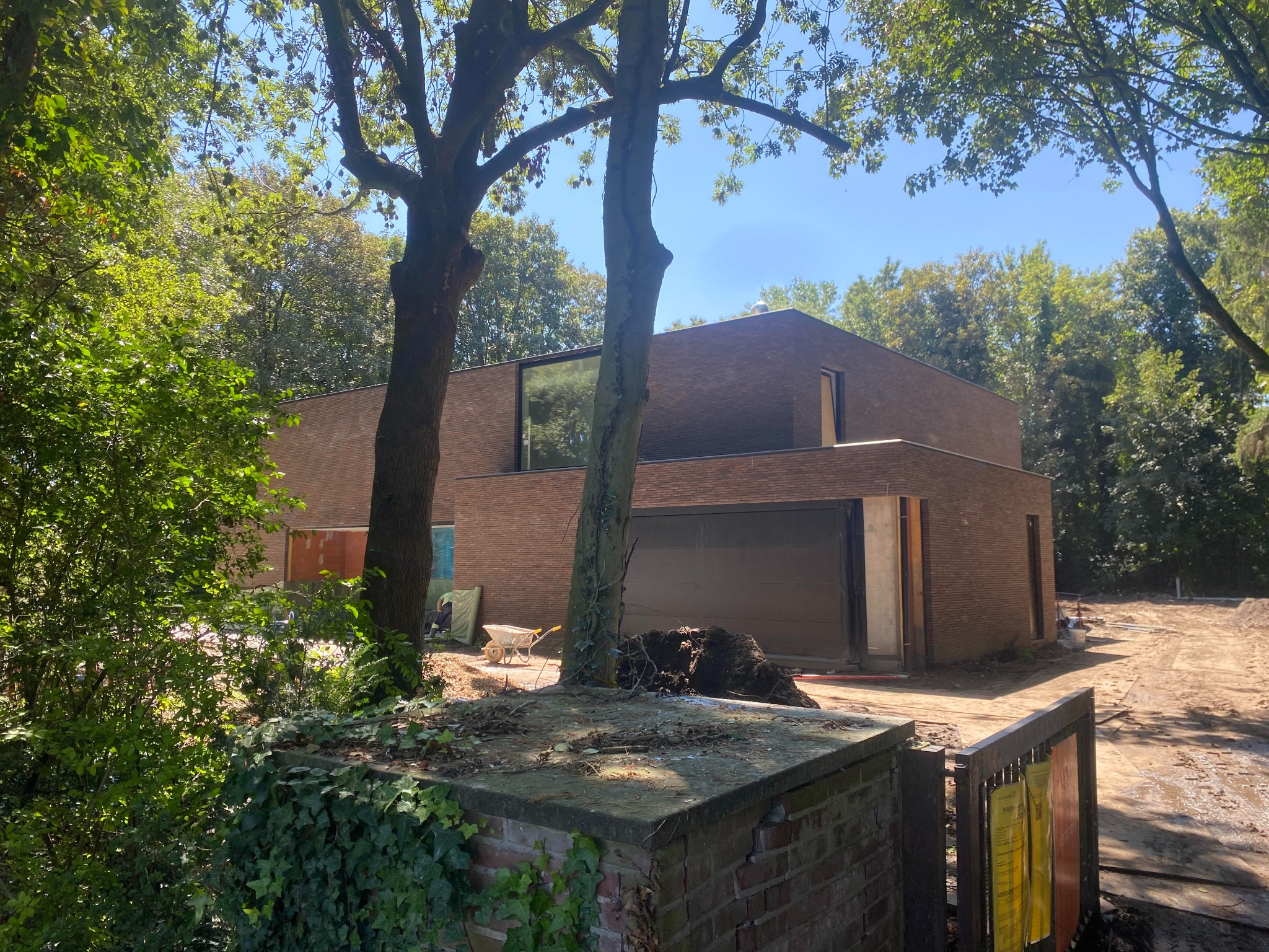Nowoczesny, jednopiętrowy dom z cegły klinkierowej w trakcie budowy, widoczny zza porośniętego bluszczem murku i drzew, z piaskiem na podwórku i elementami budowlanymi w tle.