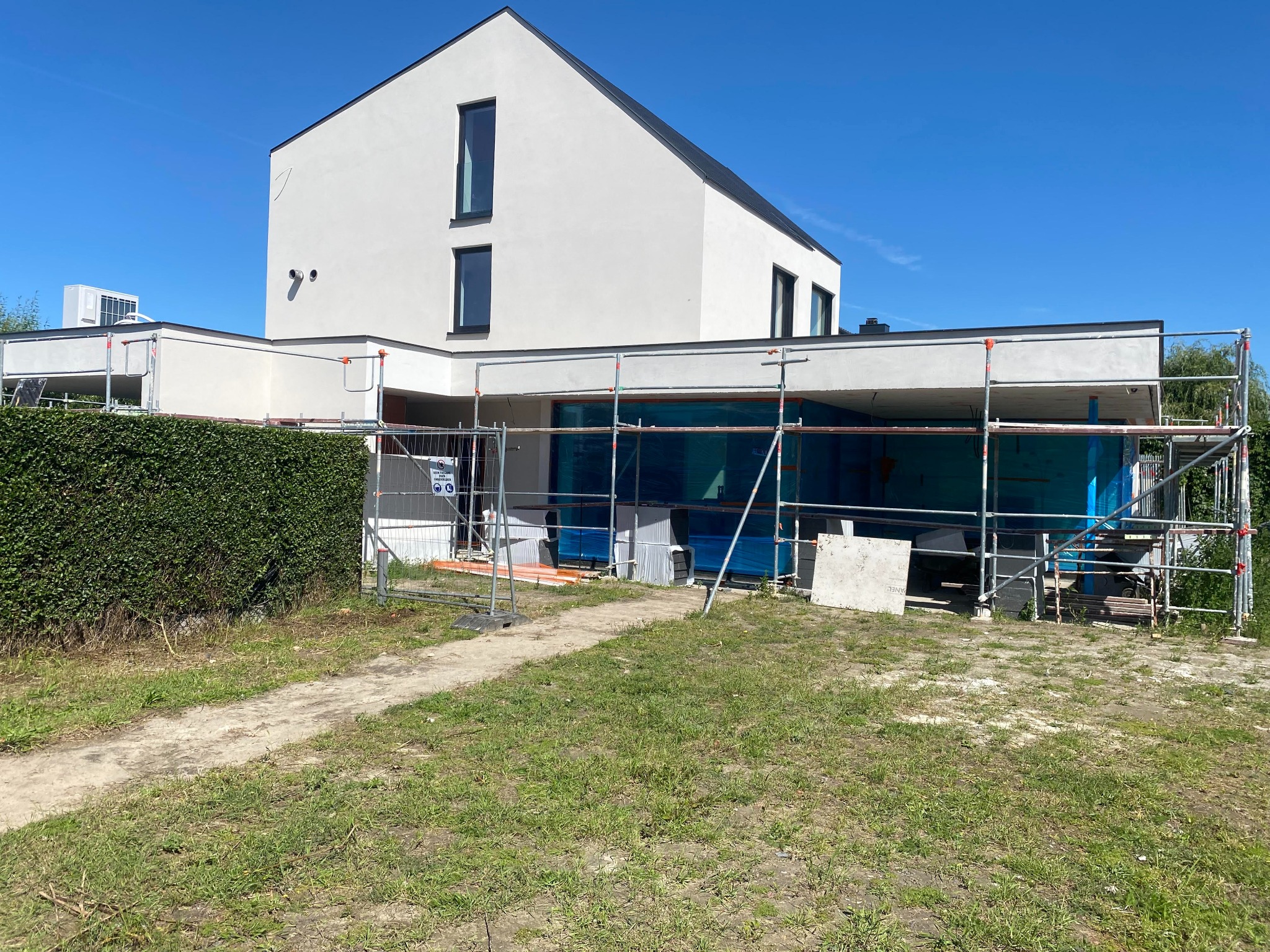 Nowoczesny dom jednorodzinny w trakcie ocieplania elewacji, widoczne rusztowania, zabezpieczone okna folią, niezagospodarowany teren wokół budynku.