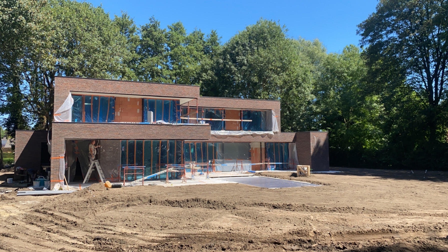 Nowoczesny dom w trakcie budowy, elewacja z cegły klinkierowej, okna zabezpieczone folią, widoczne rusztowania i prace wykończeniowe.