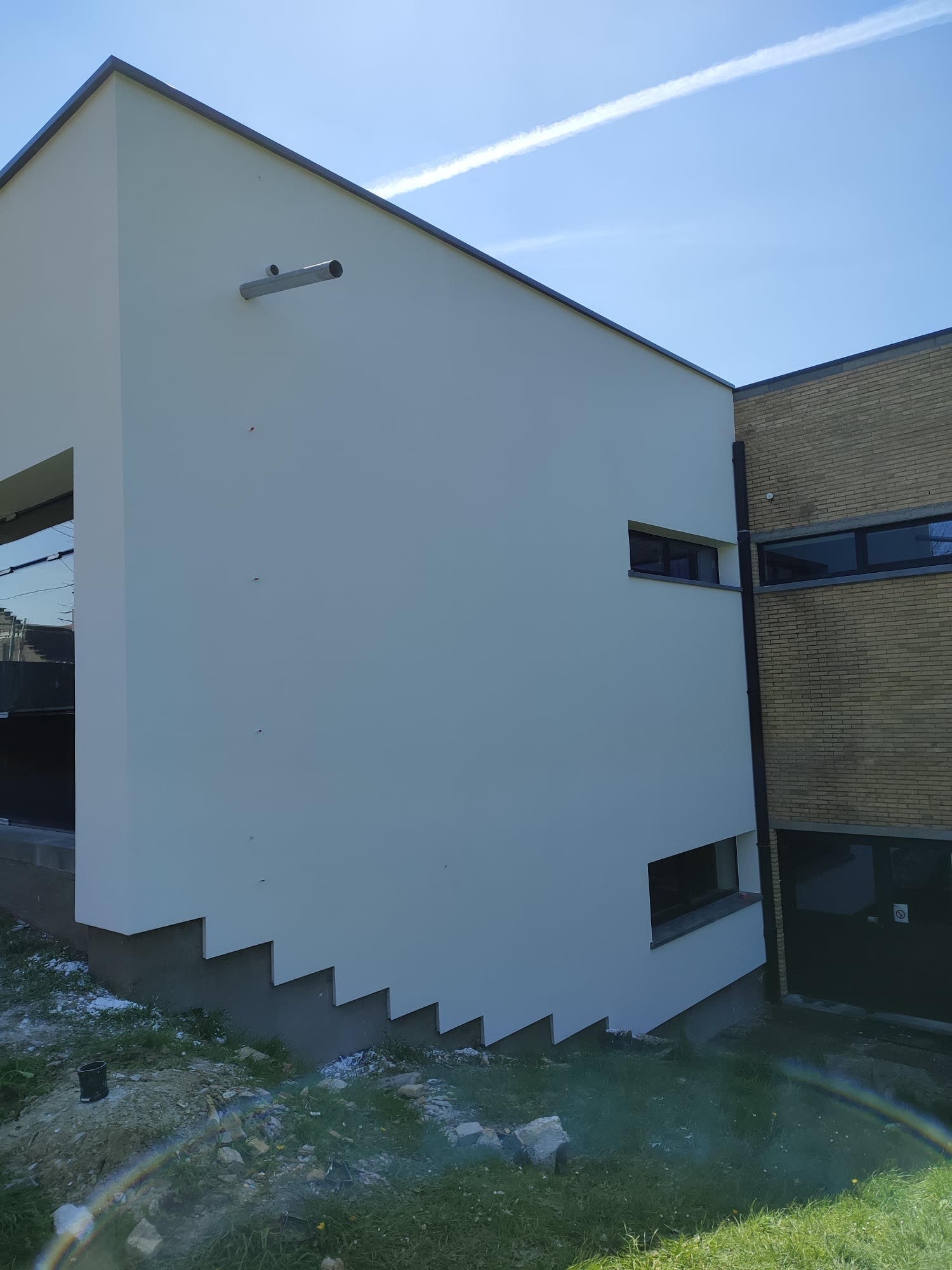 Nowoczesny budynek z białą elewacją i elementami z cegły, widoczne detale architektoniczne, takie jak schodkowa podstawa ściany i metalowa rura wentylacyjna, na tle błękitnego nieba z smugą...