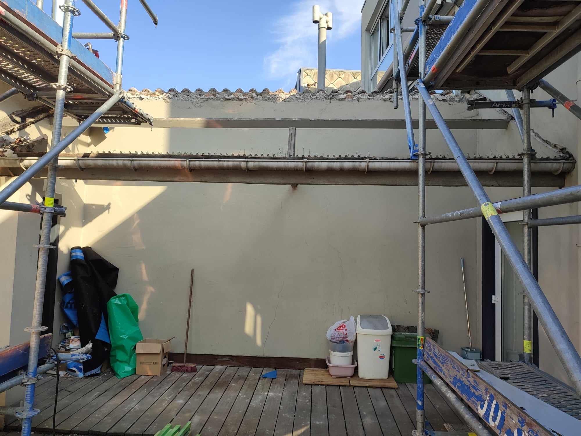 Ściana budynku w trakcie prac elewacyjnych, widoczne rusztowania, narzędzia i materiały budowlane na drewnianym podeście, fragment dachu z dachówką i rynna z kolcami przeciw ptakom.