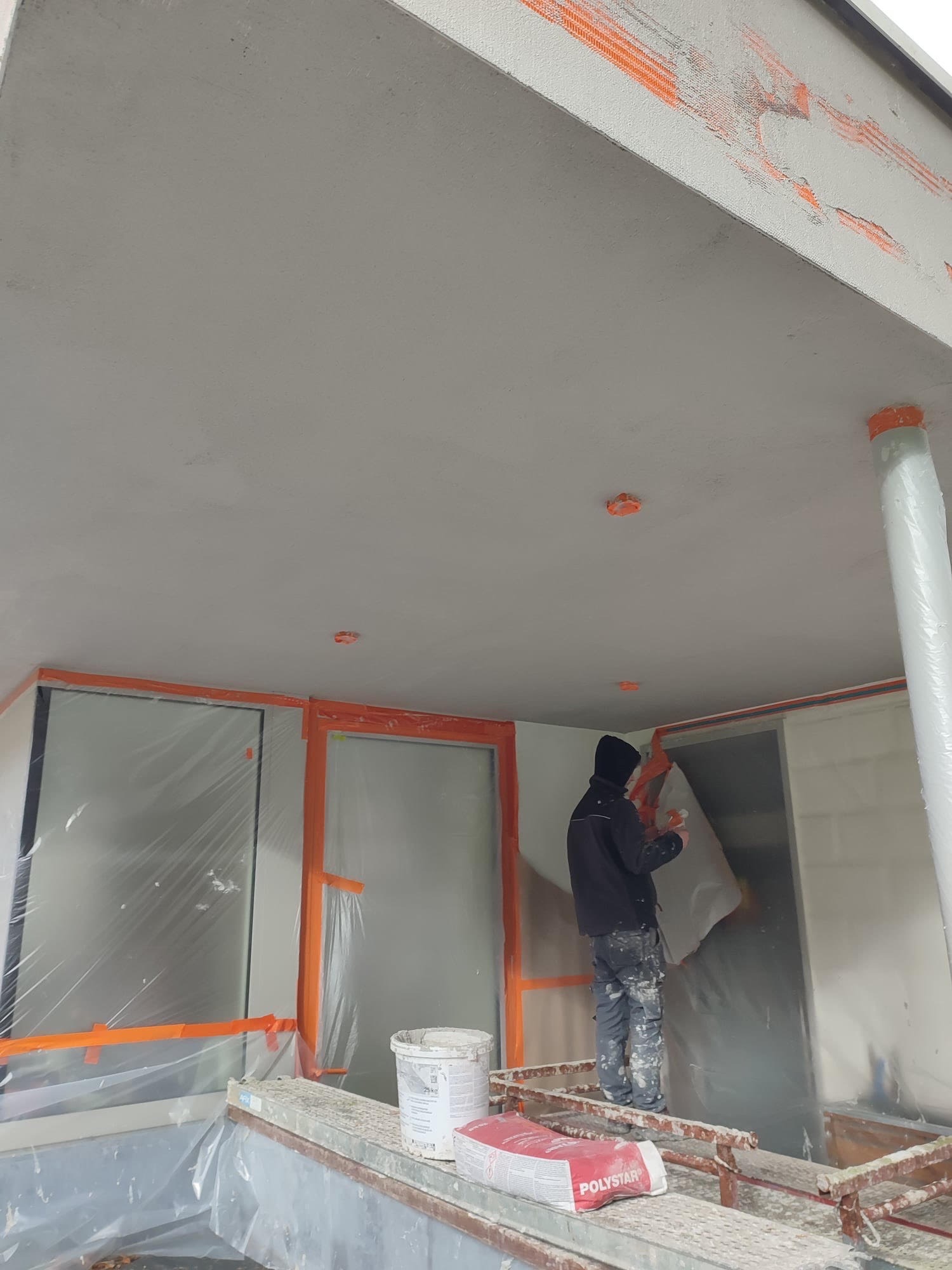 Pracownik w trakcie nakładania warstwy wykończeniowej na elewację domu jednorodzinnego, widoczne zabezpieczone okna folią i taśmą, worek z zaprawą Polystar oraz wiadro z farbą.