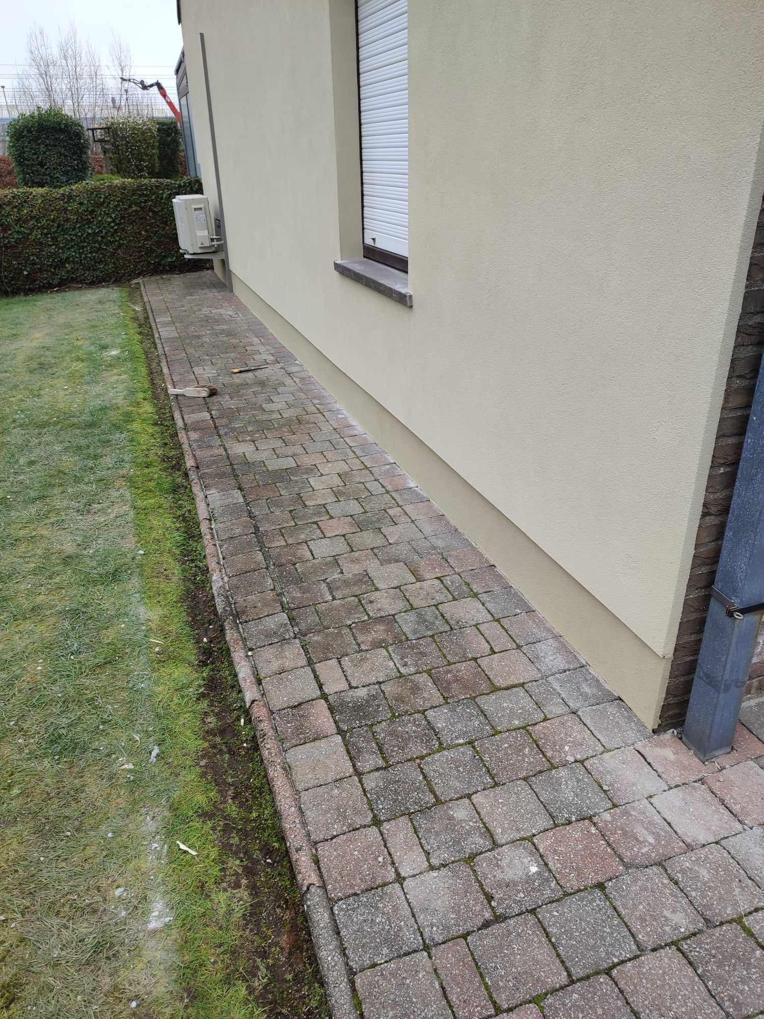 Fragment elewacji budynku z beżowym tynkiem, widoczne okno z roletą, klimatyzator na ścianie oraz chodnik z kostki brukowej. Na trawniku widoczne narzędzia.