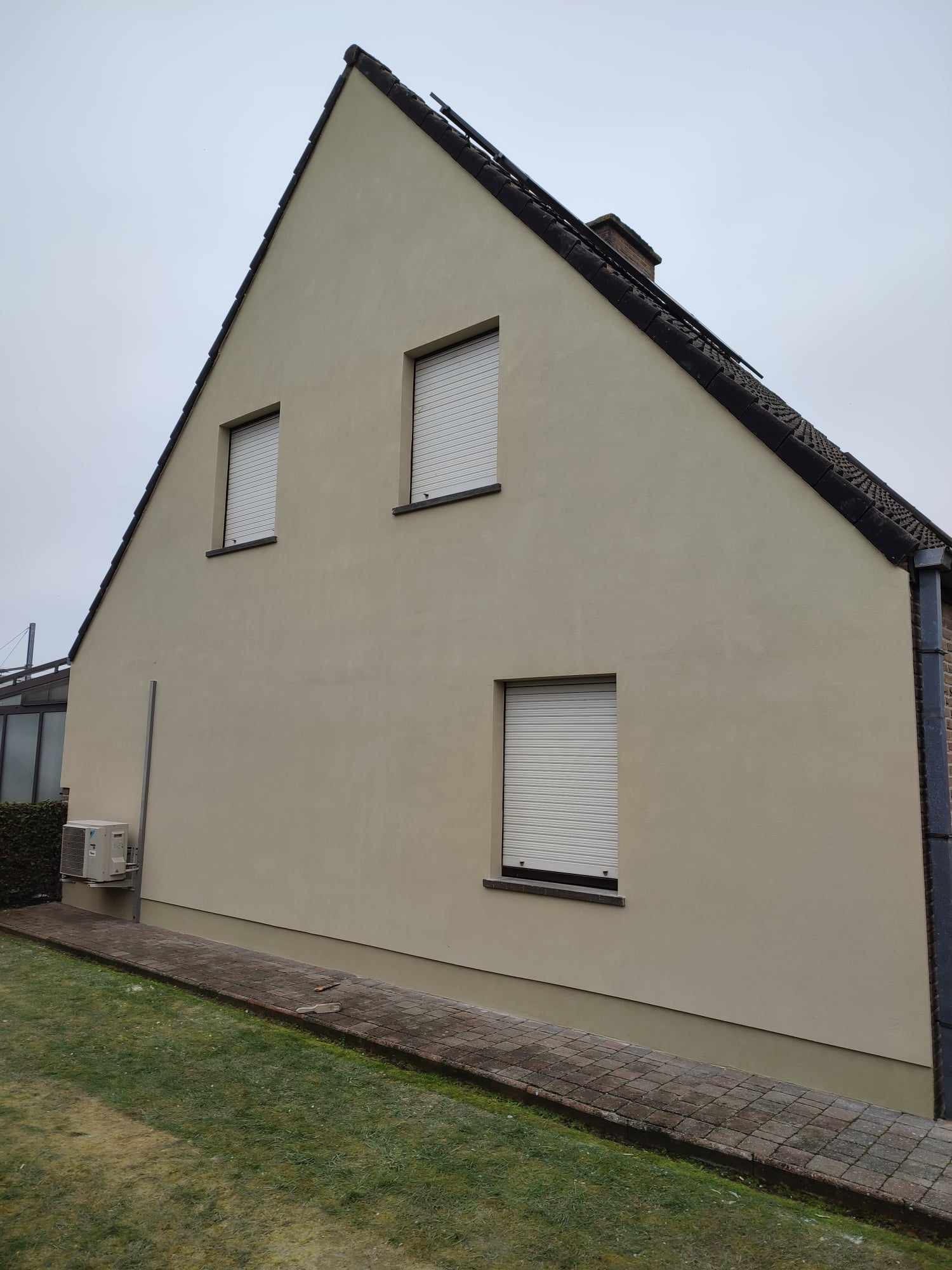 Świeżo wykończona elewacja domu jednorodzinnego z trzema oknami z roletami zewnętrznymi, widoczny fragment chodnika i trawnika oraz jednostka zewnętrzna klimatyzacji.