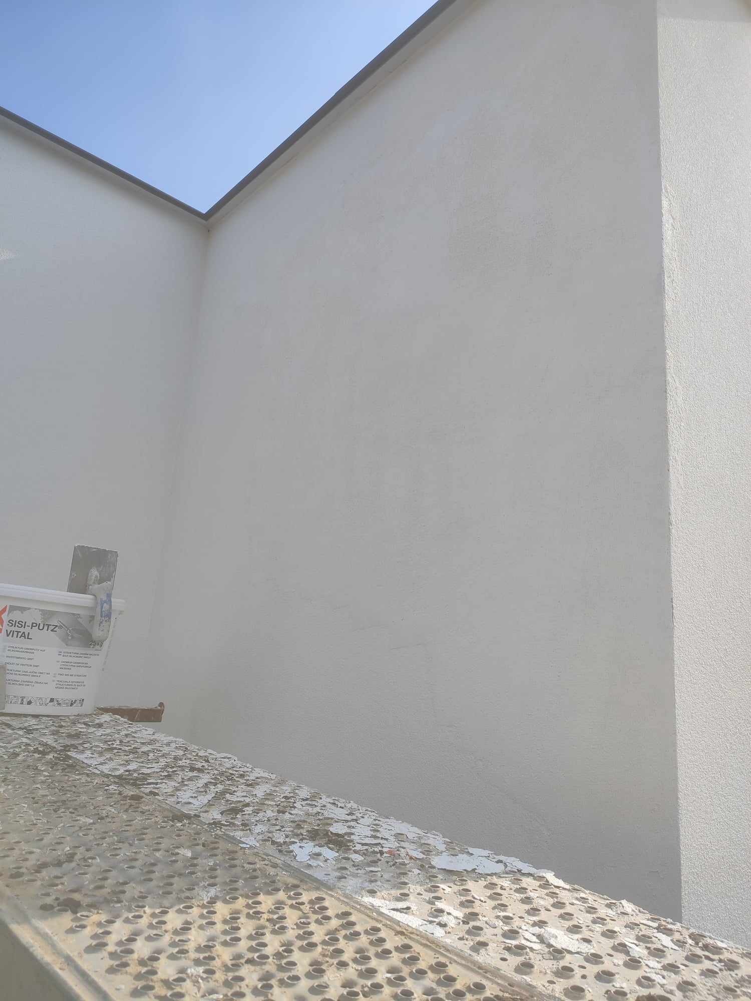Świeżo otynkowana elewacja budynku w jasnym odcieniu, widoczna z dołu, z fragmentem rusztowania i wiadrem z zaprawą SISI-PUTZ VITAL
