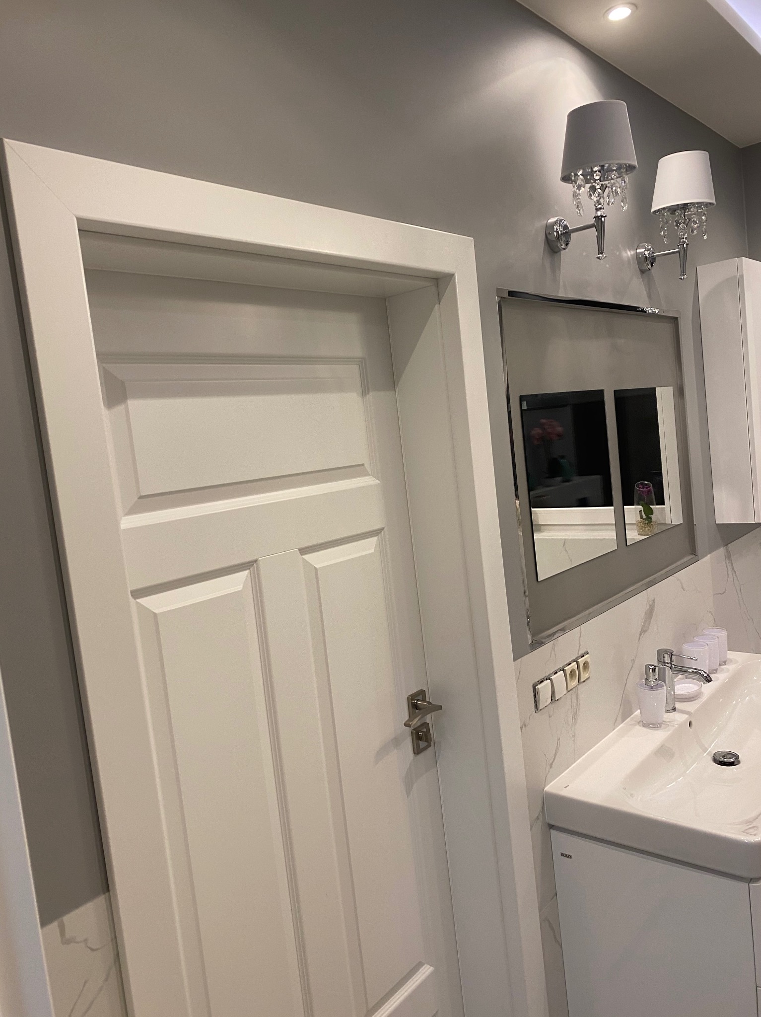 Białe, trzyskrzydłowe drzwi z ościeżnicą w łazience po remoncie, widoczna umywalka z blatem imitującym marmur, oświetlenie kinkietowe z abażurami i lustro w metalowej ramie.
