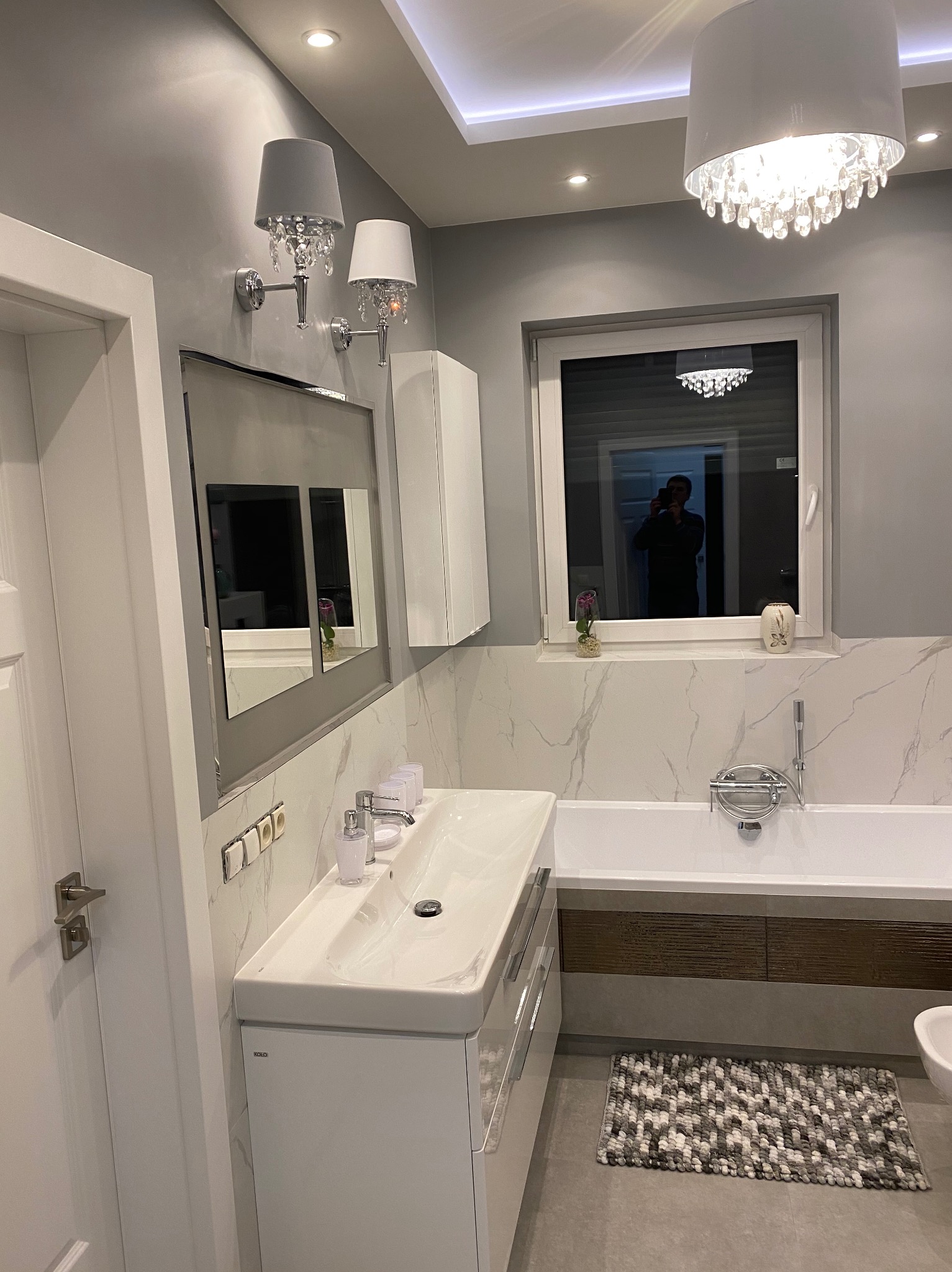 Nowoczesna łazienka w odcieniach szarości i bieli, z umywalką nablatową, lustrem w szarej ramie, kryształowymi kinkietami i dywanikiem z okrągłych kulek. W oknie widoczne odbicie osoby robiącej...