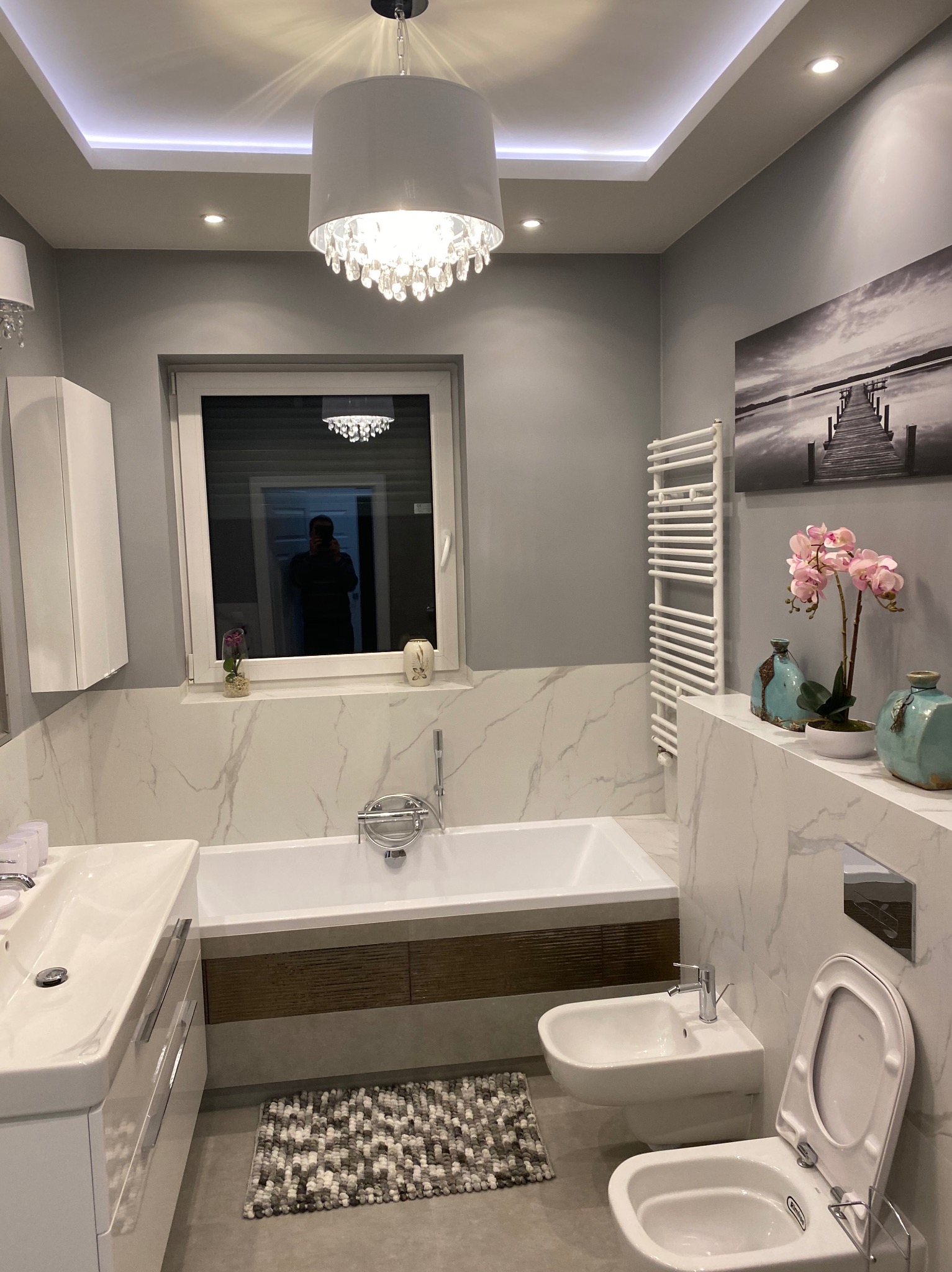 Nowoczesna łazienka w szaro-białej tonacji z marmurowymi płytkami, białą wanną z drewnopodobną obudową, wiszącą toaletą i bidetem. Nad wanną okno z odbiciem osoby, a na ścianie obraz z motywem molo.