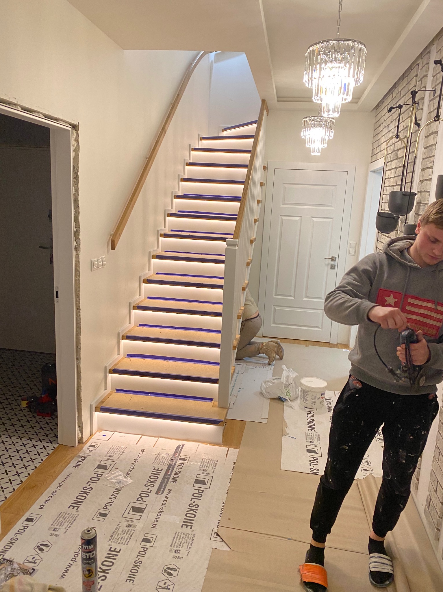 Montaż oświetlenia LED pod stopniami schodów drewnianych z białymi podstopnicami, widoczny pracownik z wiertarką, zabezpieczone podłoże folią ochronną Pol-Skone, kryształowe żyrandole w tle.