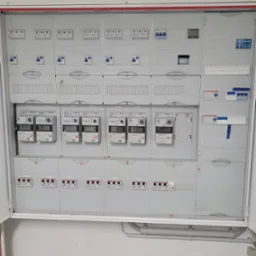 Otwarta skrzynka rozdzielcza z licznymi bezpiecznikami, wyłącznikami i licznikami energii elektrycznej, widoczne okablowanie wewnątrz, szara kolorystyka.