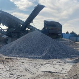 Sort Krusz - Góra drobnego kruszywa wysypana z maszyny przesiewającej, obok hałda piasku, widoczne ślady opon i budynki w tle.