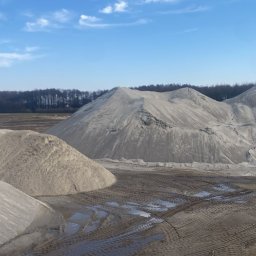 Sort Krusz - Kopalnia odkrywkowa z hałdami jasnego kruszywa, widoczne ślady opon i kałuże, w tle drzewa pod błękitnym niebem.