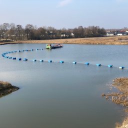 Sort Krusz - Pogłębianie dna zbiornika wodnego przy użyciu pływającej pogłębiarki z rurociągiem transportującym urobek, widoczne brzegi zbiornika z roślinnością i zabudowania w oddali.