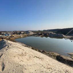 Sort Krusz - Kopalnia odkrywkowa z jeziorem w centrum, hałdy materiału, ciężki sprzęt budowlany w tle, błękitne niebo.