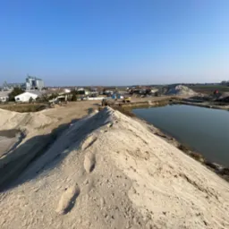 Widok z góry na hałdę materiału sypkiego z widocznymi śladami, obok zbiornik wodny, w tle zabudowania przemysłowe i sprzęt budowlany.