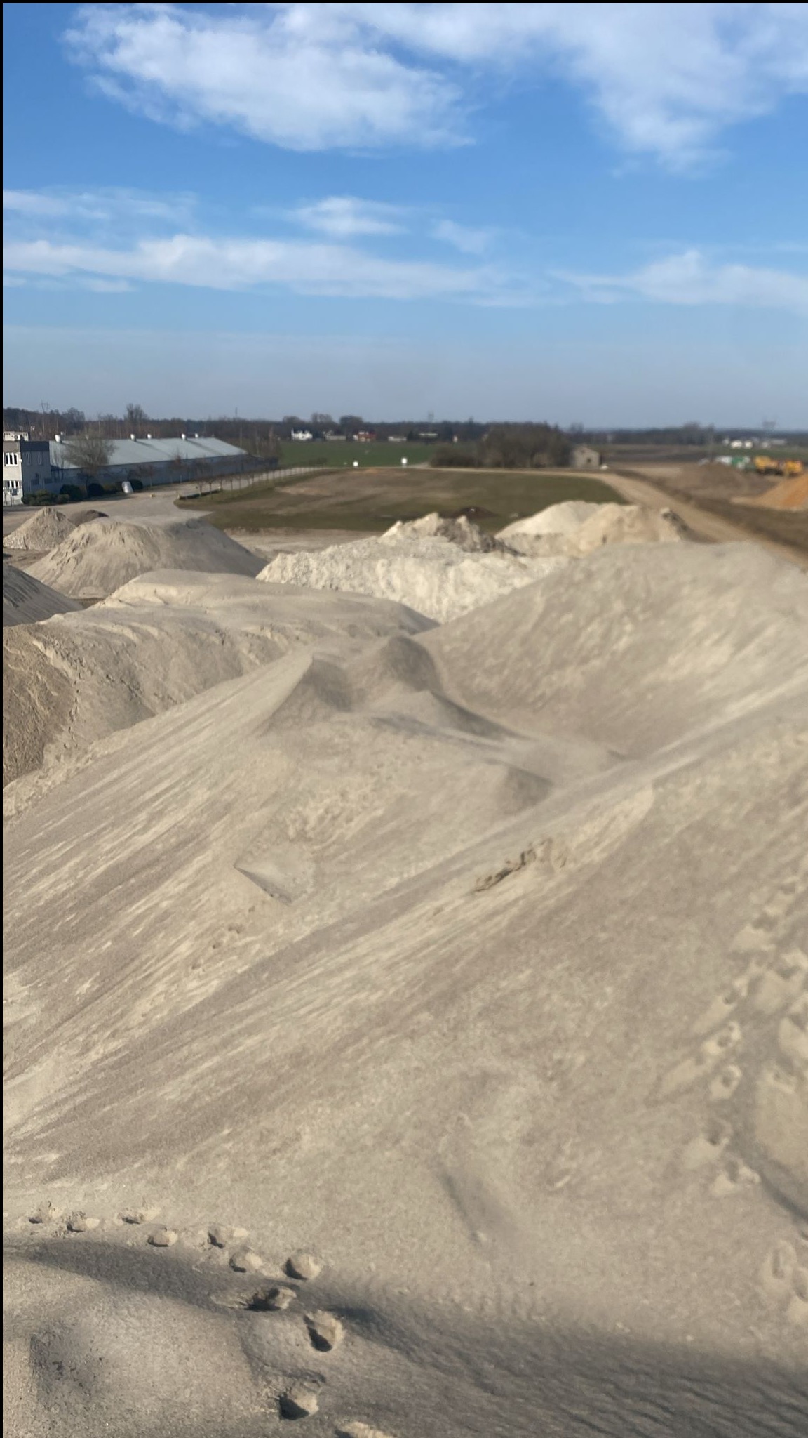 Góra piasku z widocznymi śladami stóp w Mierzynku, na tle błękitnego nieba z chmurami i budynków w oddali.